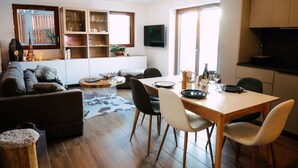 Uma sala de estar moderna com um sofá, uma mesa de centro e uma mesa de jantar posta para quatro pessoas. Há estantes embutidas e uma TV de tela plana fixada na parede.