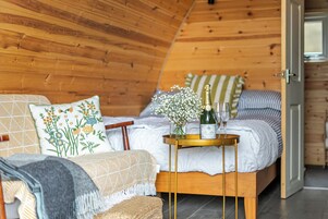 Un dormitorio acogedor con techo de madera, cama con colcha blanca y estampada, mesita de noche con una botella de champán y un florero con flores.