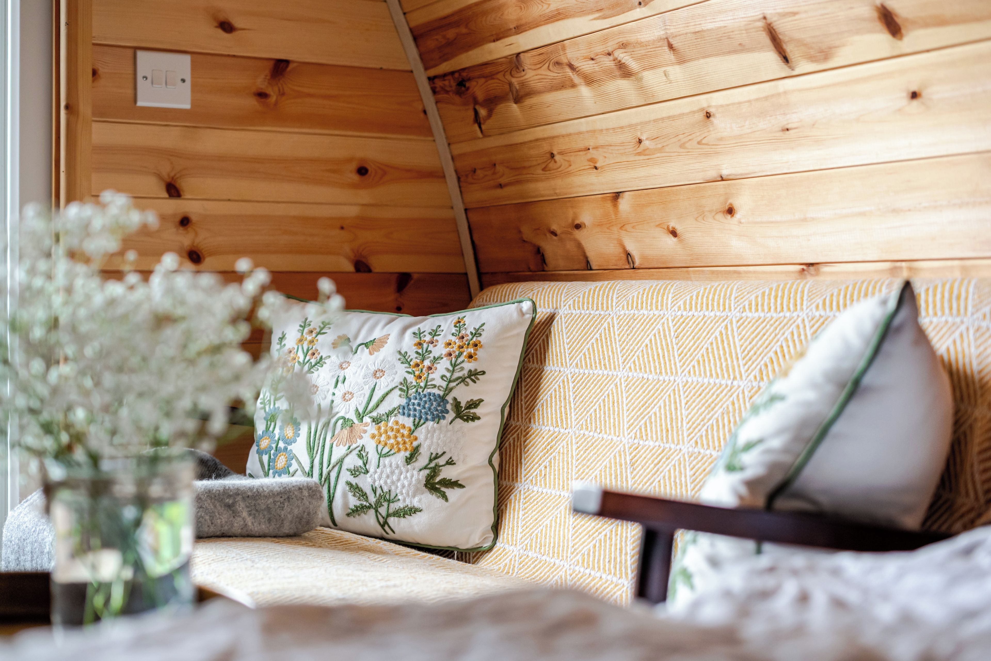 A wooden interior with a sofa and cushions, a wall switch, and a vase with flowers.