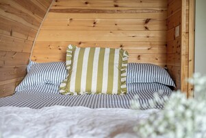 Interior de una cabaña de madera con una cama, ropa de cama a rayas y un almohadón de rayas verticales.