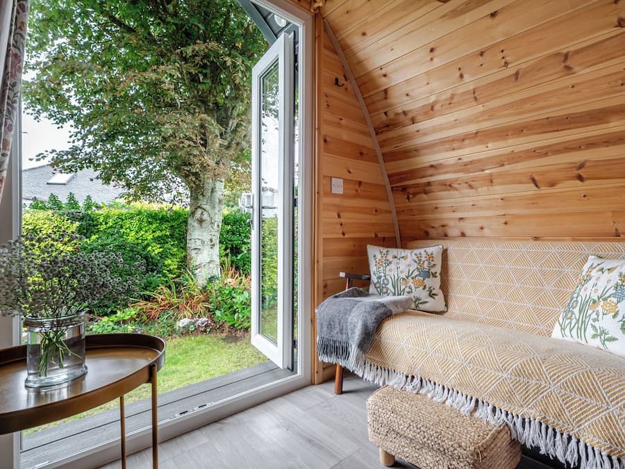 A cozy room with a wooden ceiling, a sofa with patterned cushions, a small table with a vase, and a view of greenery outside.