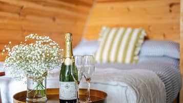 A bottle of champagne, two glasses, and a vase with flowers on a tray in a room with wooden walls and a bed with striped pillows.