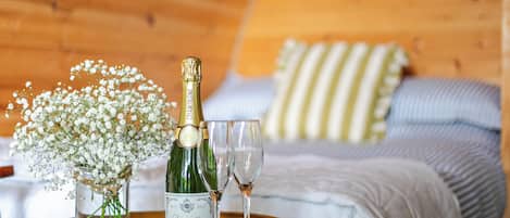 A bottle of champagne, two glasses, and a vase with flowers on a tray in a room with wooden walls and a bed with striped pillows.