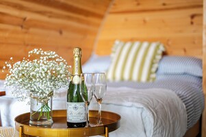 Una botella de champán, dos copas y un florero con flores sobre una bandeja en una habitación con paredes de madera y una cama con almohadones a rayas.