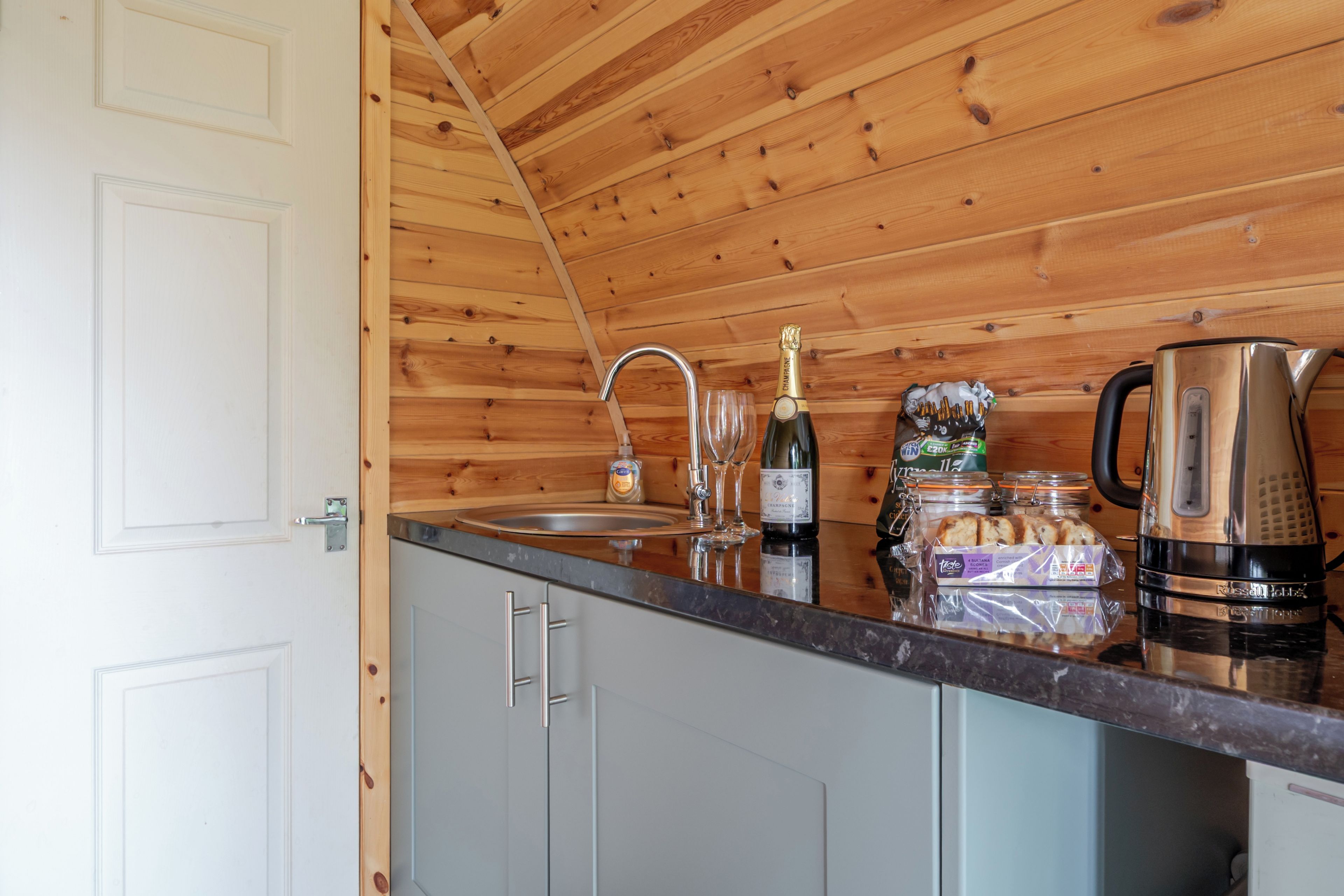 A kitchen with wooden walls, a stainless steel sink, a wine bottle, and a kettle.