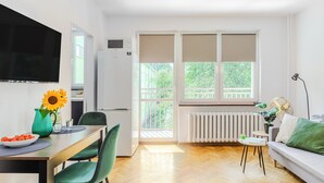 Ein modernes Wohnzimmer mit einer Couch, einem Esstisch mit Stühlen, einem an der Wand montierten Fernseher und einem großen Fenster mit Jalousien.
