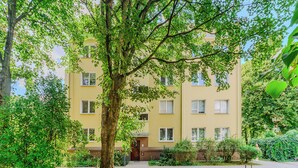Ein mehrstöckiges, gelbes Gebäude mit einem zentralen Eingang, umgeben von üppiger Vegetation und Bäumen.