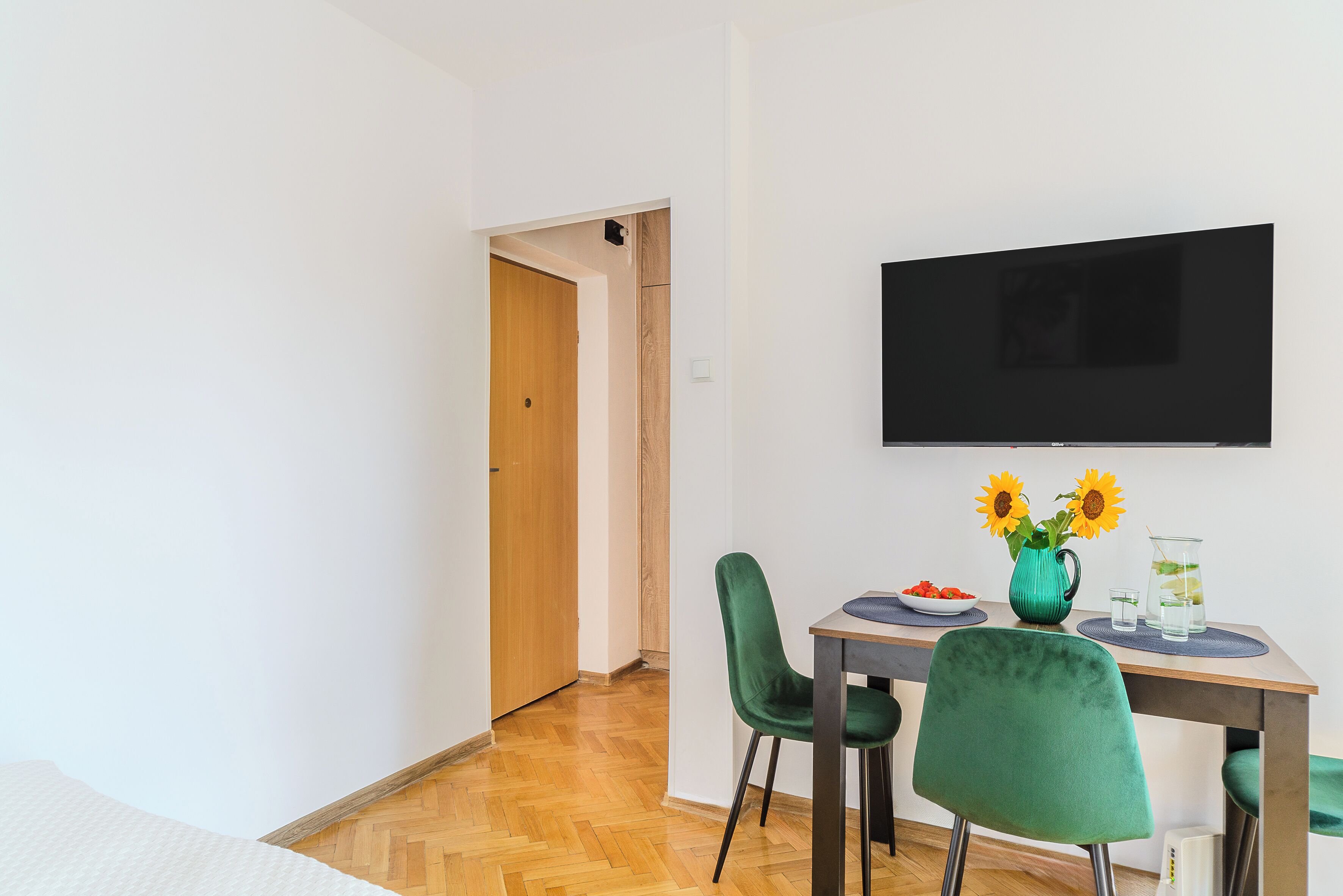 A modern room with a wooden floor, a dining table with chairs, a flat-screen TV, and a vase with sunflowers.
