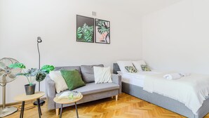 Ein modernes Zimmer mit grauem Sofa, einem kleinen Tisch mit Ventilator und Pflanze, einem Bett mit weißer Bettwäsche sowie zwei an der Wand hängenden Kunstwerken.
