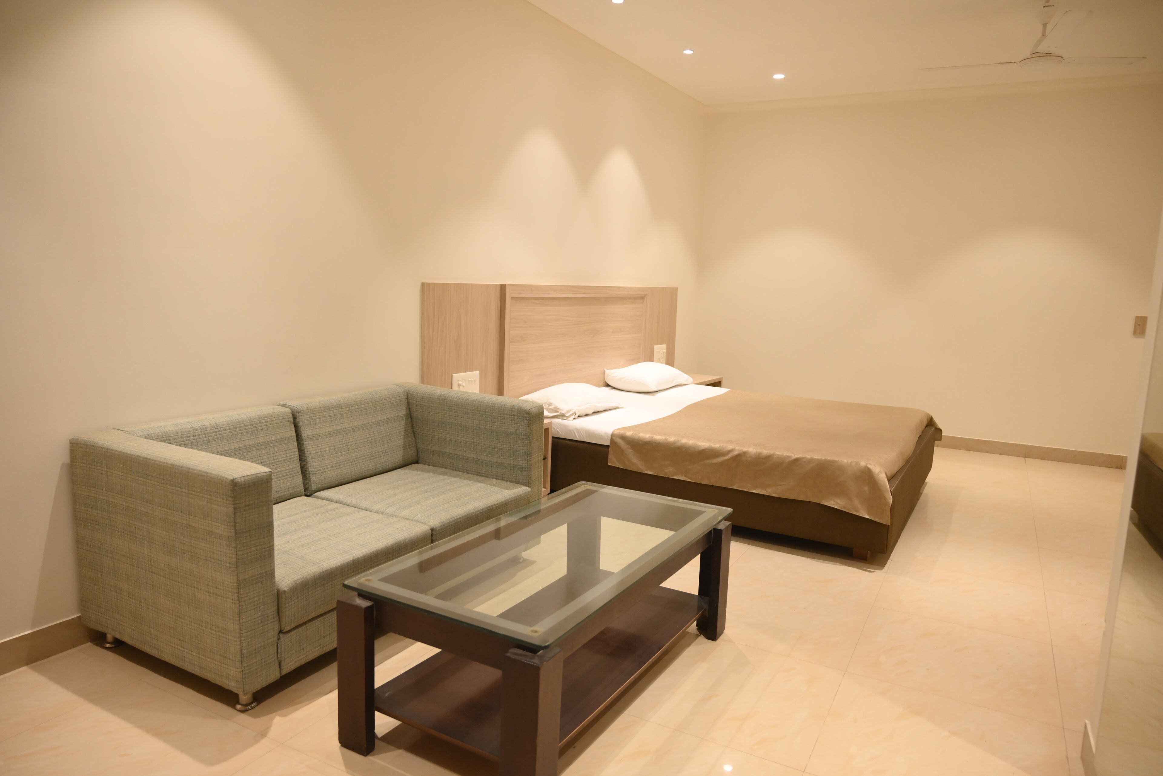 Una habitación de hotel moderna con un sofá verde, una mesa de centro de vidrio, una cama con una manta color crema y un cabecero de madera.