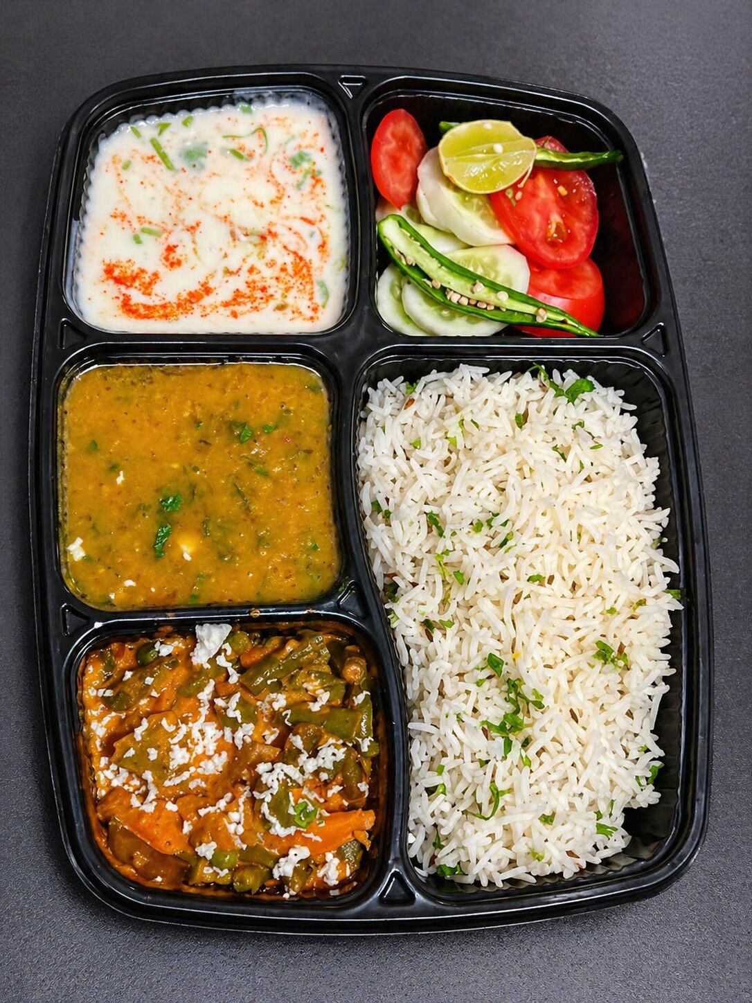 A meal in a black compartmentalized tray with compartments for rice, a vegetable curry, a lentil dish, and a yogurt-based dish.