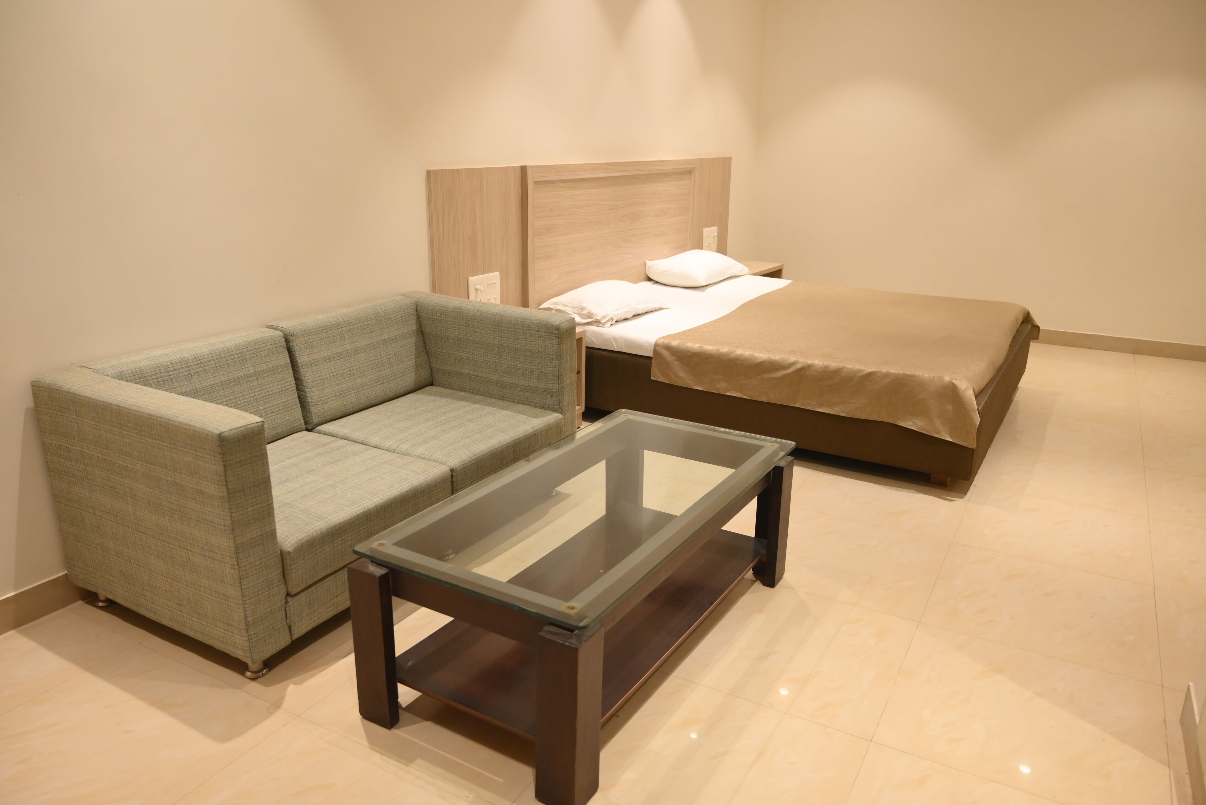 A hotel room with a green sofa, a glass-top coffee table, a bed with a brown cover, and a wooden headboard.