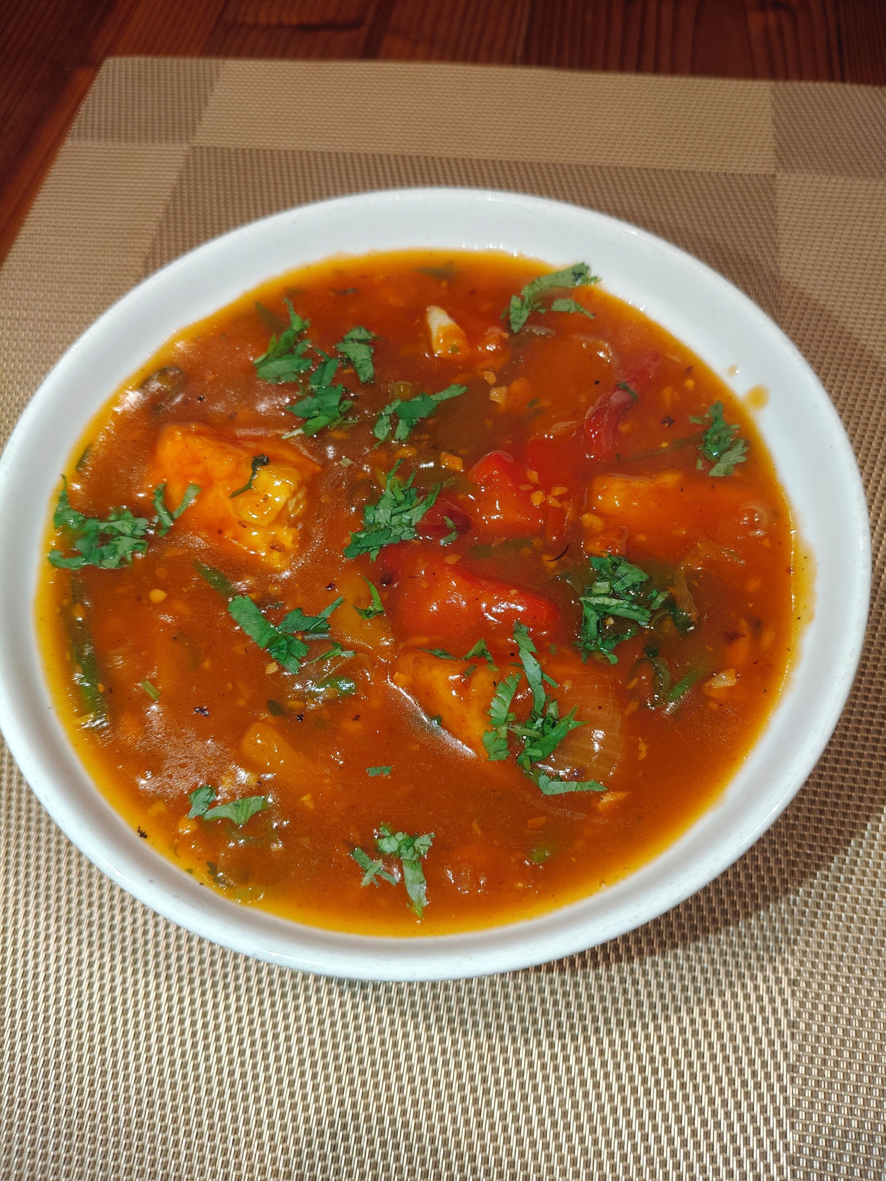 A bowl of soup with visible ingredients like tomatoes and herbs, placed on a textured mat.