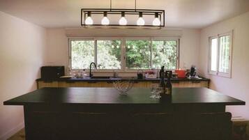 Une cuisine dotée d’un grand îlot central, d’une fenêtre donnant sur la verdure et d’un luminaire moderne placé au-dessus de l’îlot.