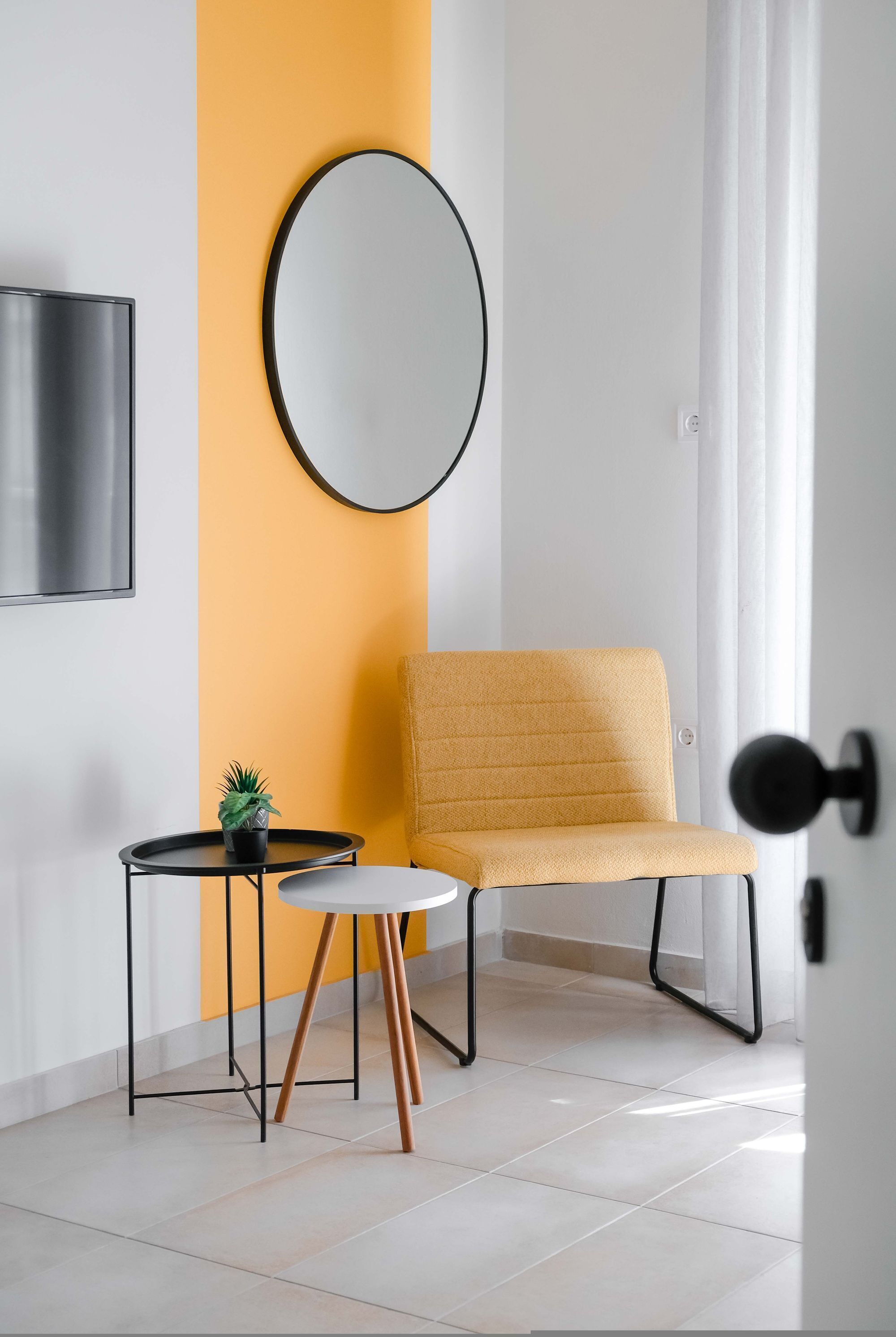 A modern room with a yellow accent wall, a round mirror, a mustard-colored chair, a small round table with a plant, and a door with a black handle.