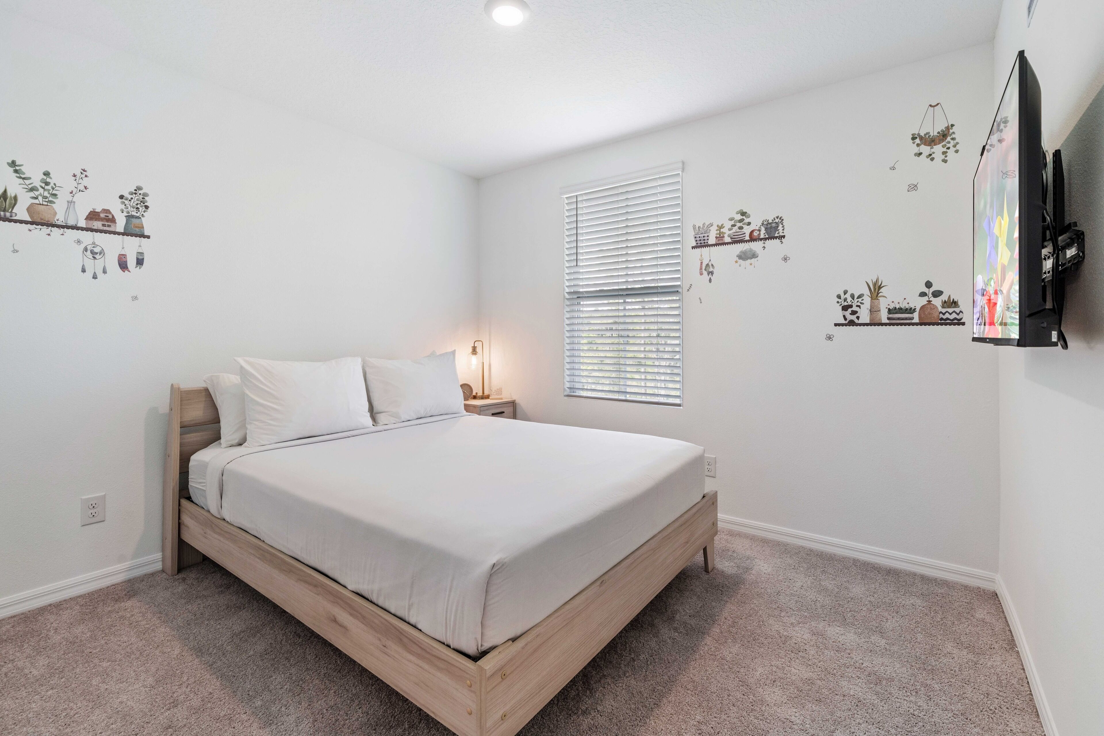 A bedroom with a wooden bed, white bedding, a nightstand, a lamp, a window with blinds, and wall decorations including plants and a TV.
