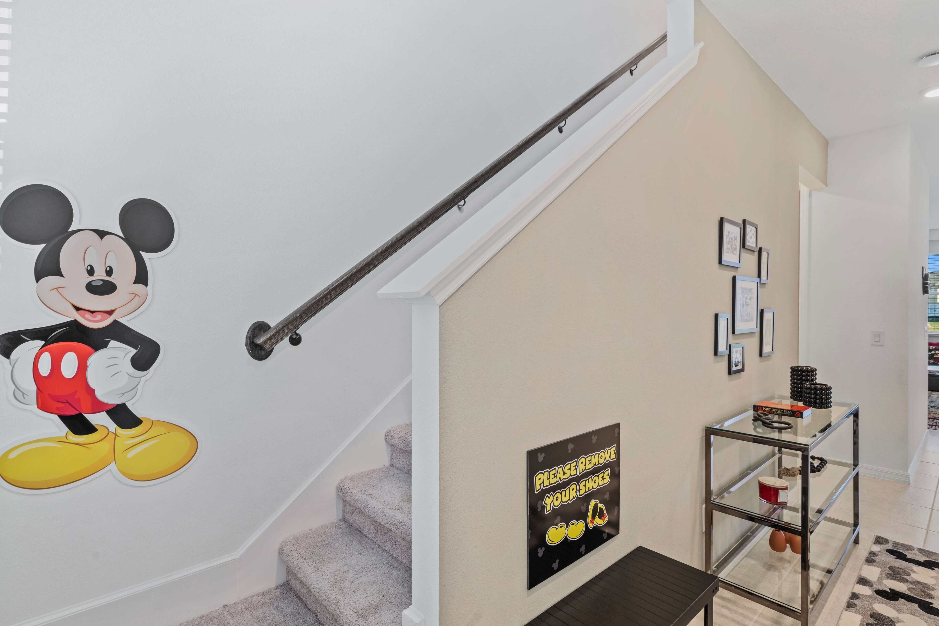 A staircase with a Mickey Mouse decal, a sign instructing to remove shoes, and a glass shelf with various items.