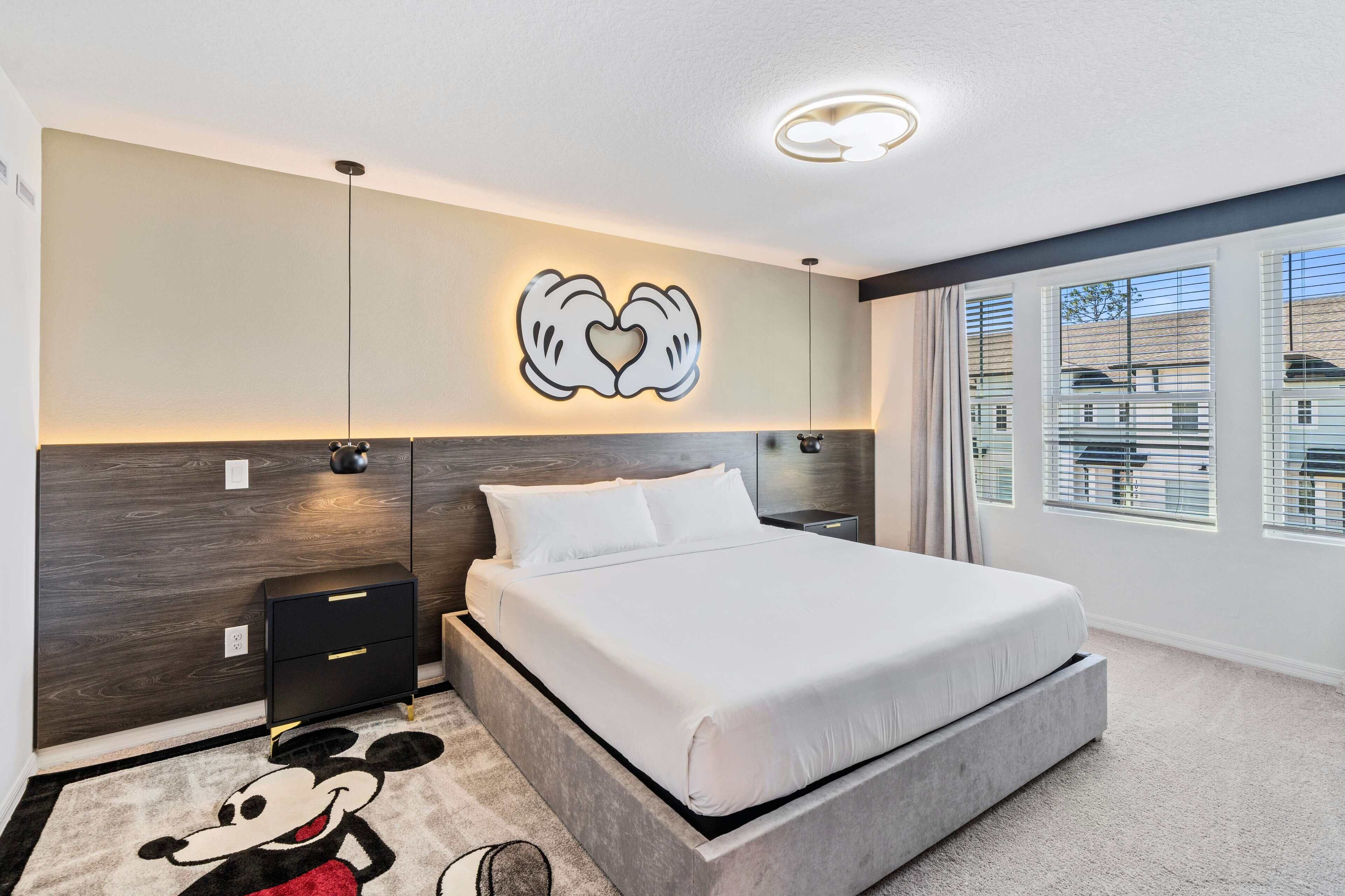 Une chambre moderne avec un grand lit, un tapis à l’effigie de Mickey Mouse, une décoration murale représentant deux cœurs et une vue sur l’extérieur grâce à de grandes fenêtres.