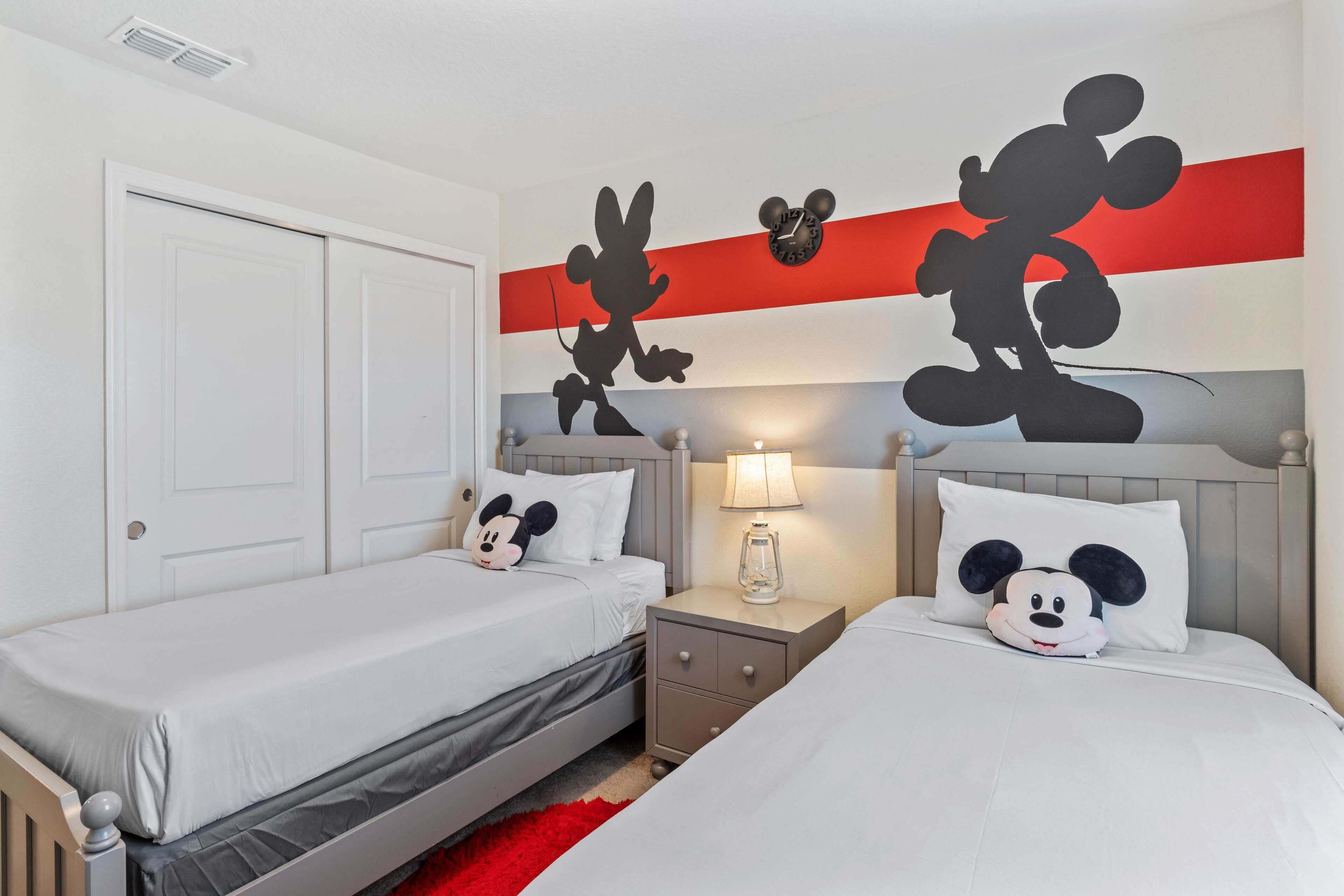 A hotel room with two beds, each with Mickey Mouse pillows, a nightstand with a lamp, and a red accent wall.
