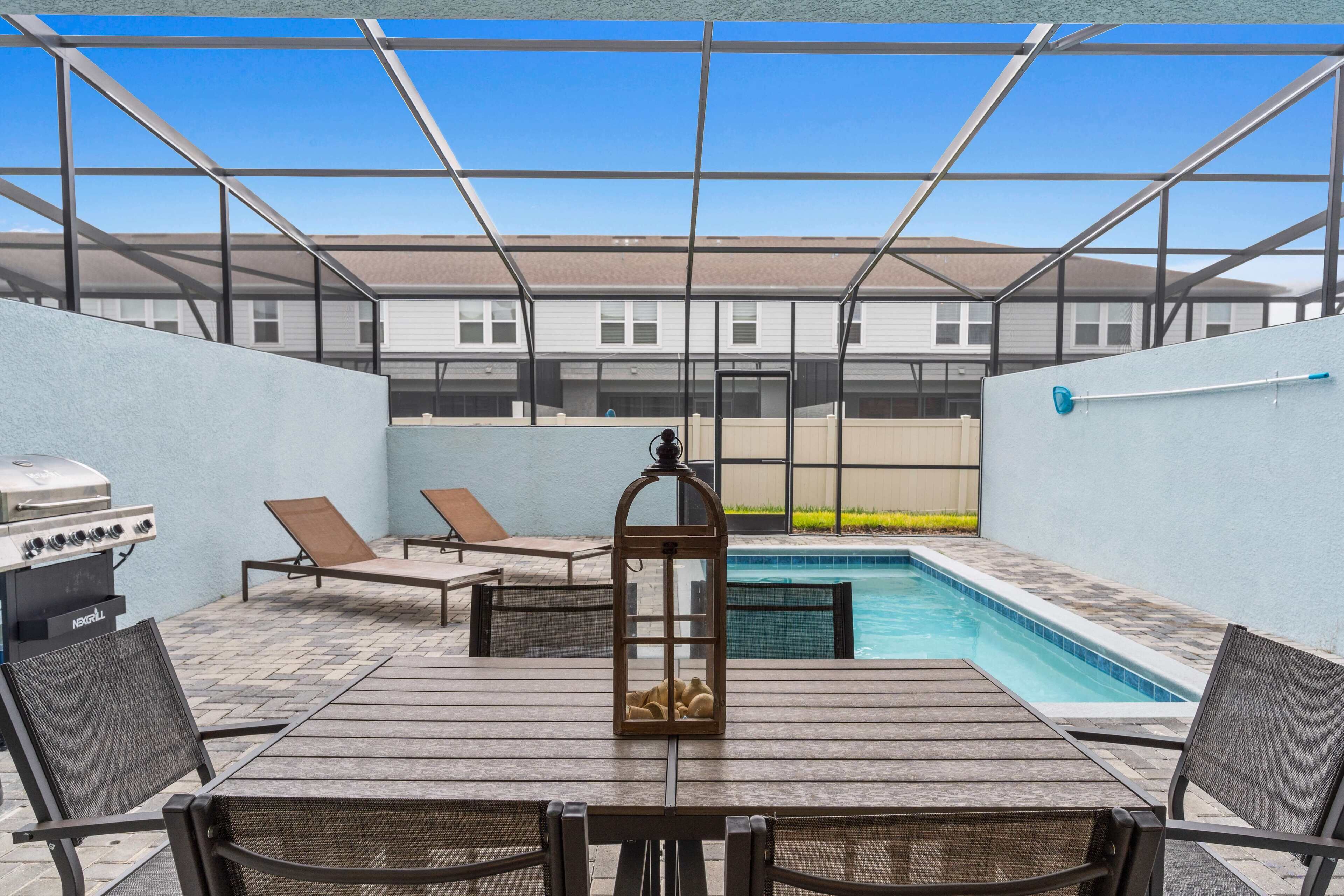 An outdoor patio with a pool, lounge chairs, a grill, and a table.