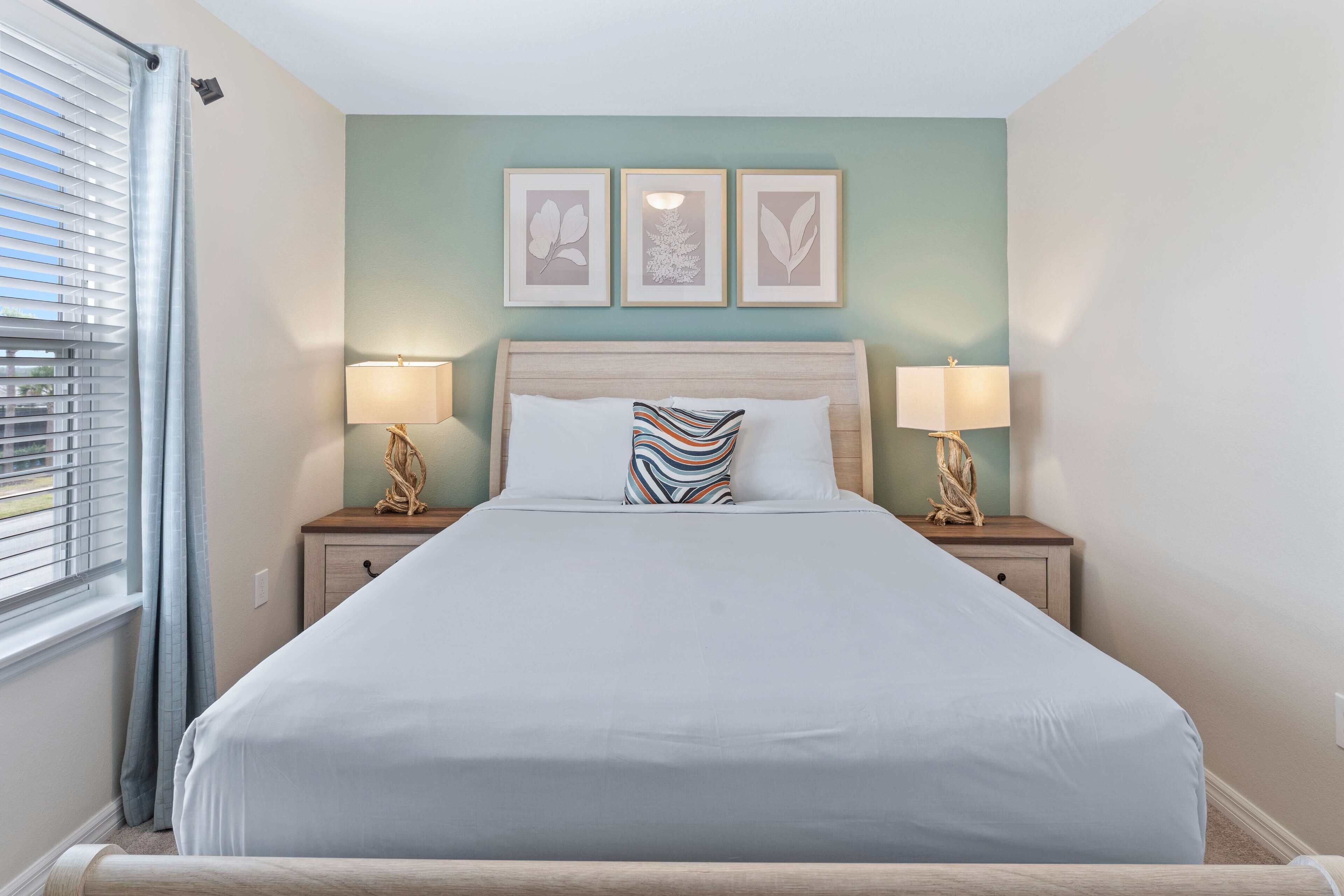 A neatly made bed with a white comforter and striped pillow, flanked by two bedside tables with lamps, in a room with a light green wall and three framed botanical prints above the bed.