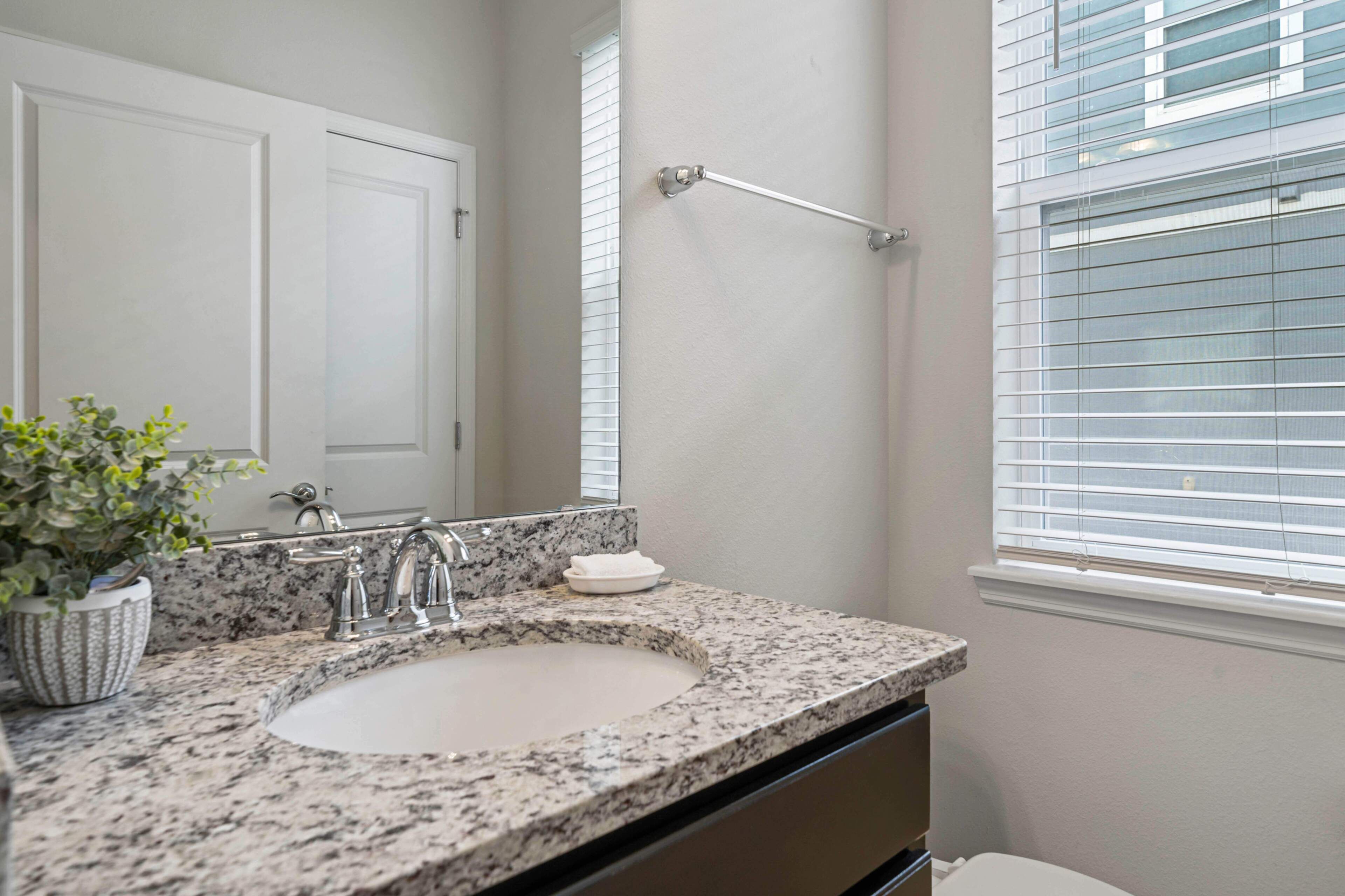 A bathroom with a granite countertop sink, a mirror, a towel rack, and a window with blinds.