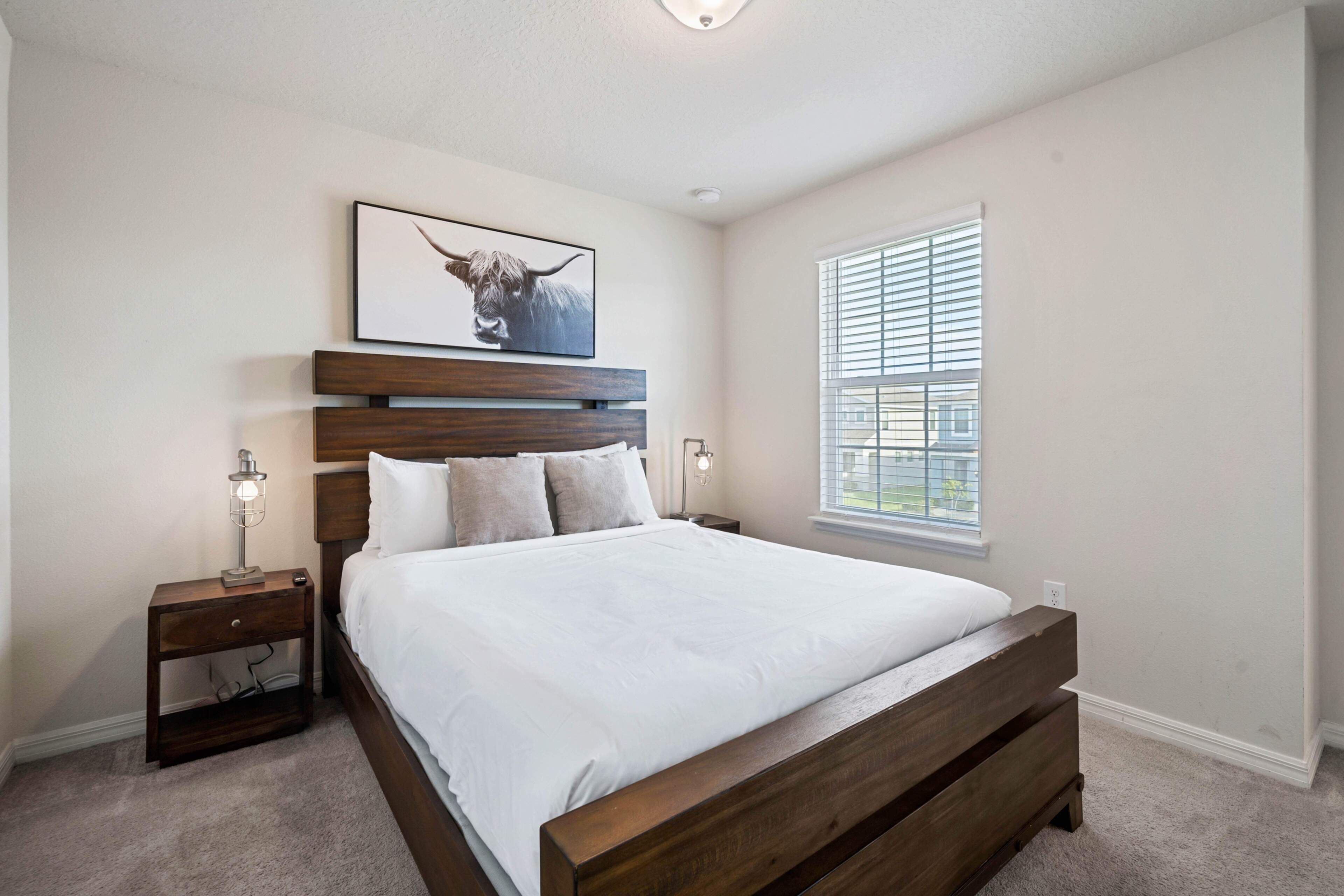 A bedroom with a wooden bed, a nightstand with a lamp, a framed picture on the wall, and a window with blinds.