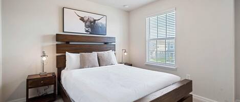 A bedroom with a wooden bed, a nightstand with a lamp, a framed picture on the wall, and a window with blinds.