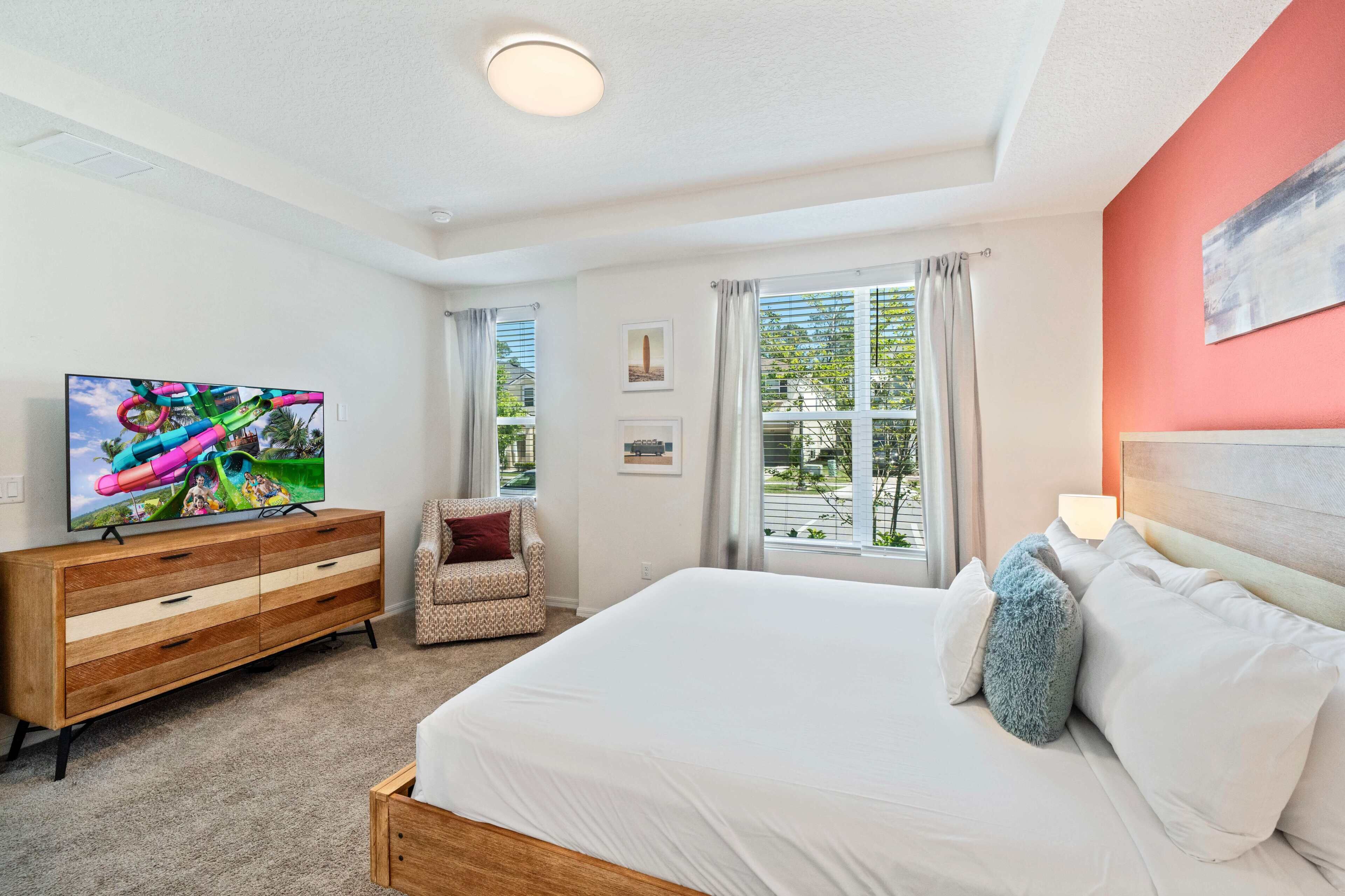 Une chambre à coucher avec un lit, une télévision, une commode en bois, une chaise et une fenêtre avec des rideaux.