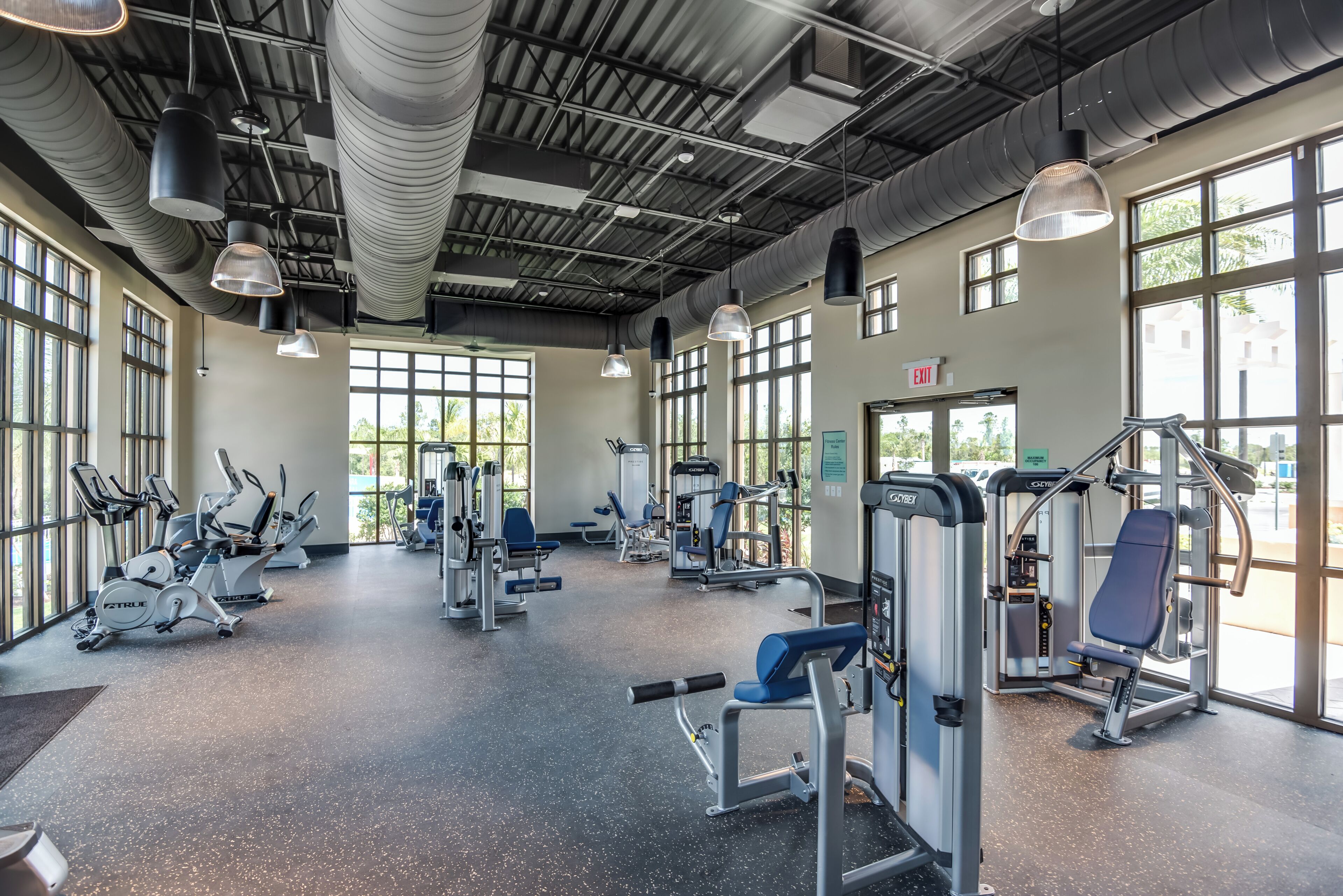 A spacious gym with various exercise machines, large windows, and a high ceiling with industrial-style lighting.