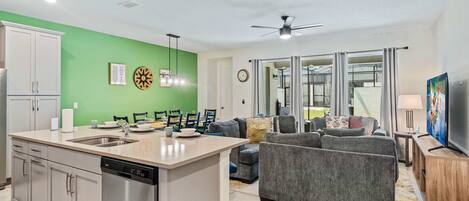 A modern kitchen with a dishwasher, a dining area with chairs and a table, a grey sofa, and a TV.