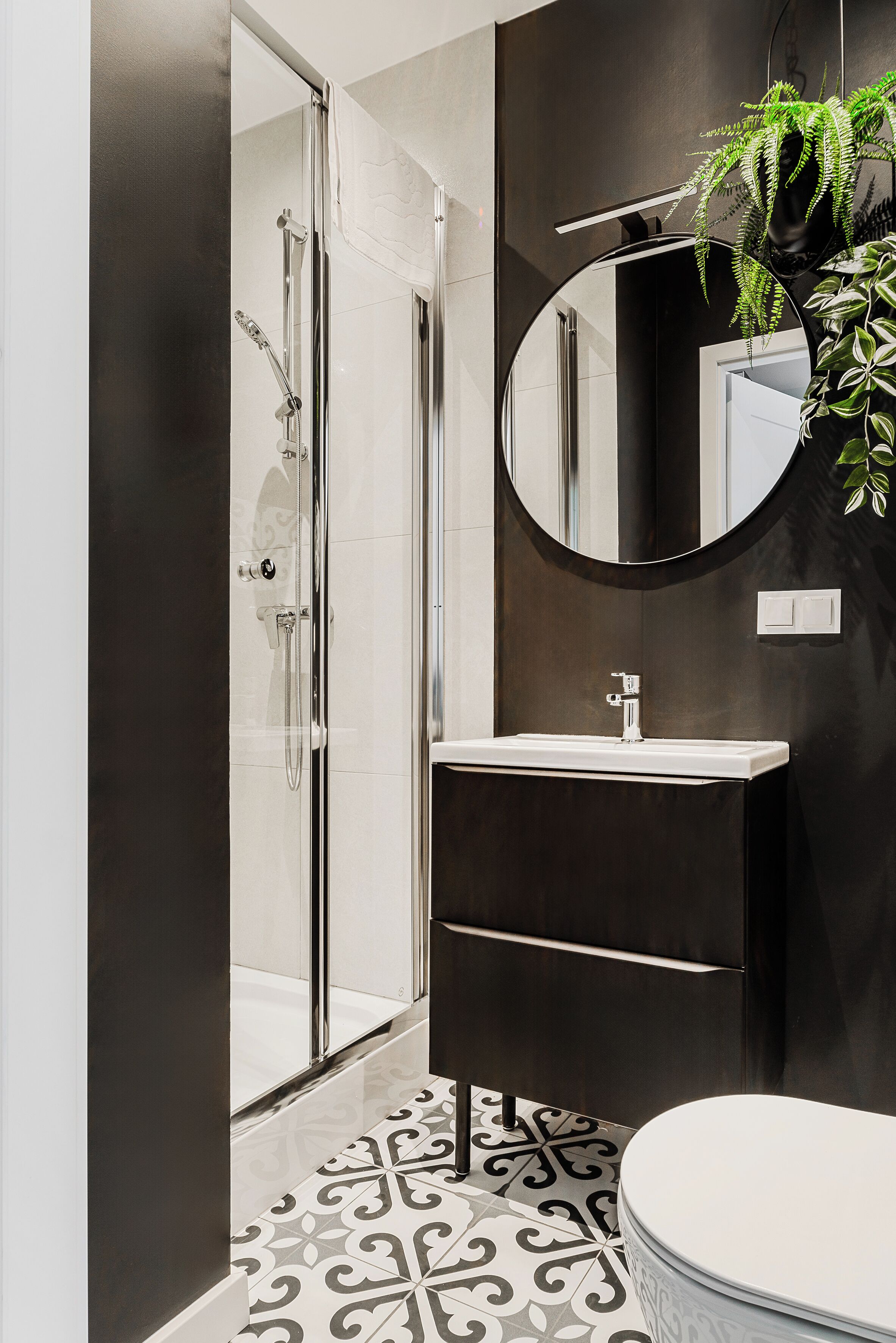A modern bathroom with a glass shower enclosure, a round mirror, a sink, and a toilet.