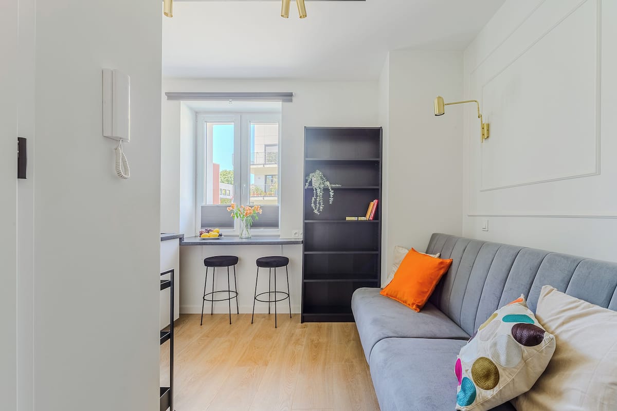 Ein modernes Wohnzimmer mit grauem Sofa, orangefarbenem Kissen, einem schwarzen Bücherregal und einer kleinen Küchenzeile mit Barhocker.