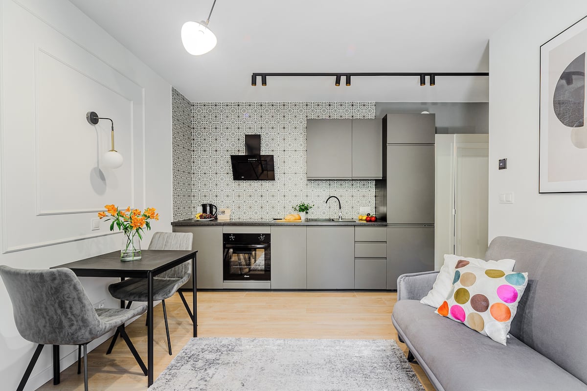 Eine moderne Küche mit grauen Schränken, einer schwarzen Dunstabzugshaube und einem eingebauten Backofen. Es gibt einen Essbereich mit Tisch und Stühlen sowie ein graues Sofa mit farbigem Kissen.