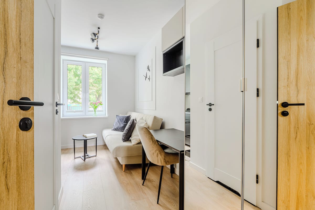 Ein modernes, helles Zimmer mit einer Holztür, einem weißen Sofa, einem kleinen runden Tisch und einem Essbereich mit dunklem Tisch und Stühlen.