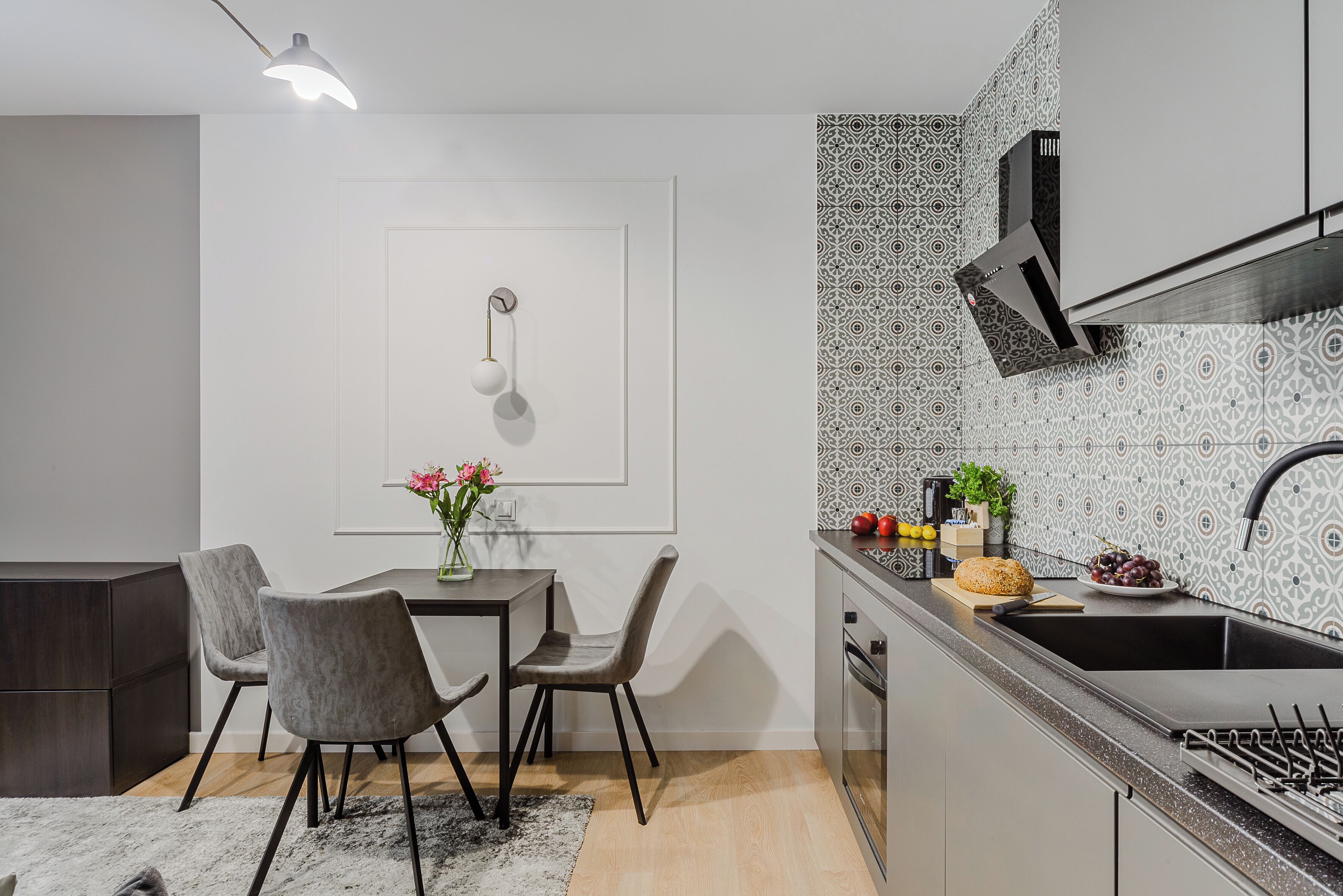 A modern kitchen with a dining area, featuring a dark wood cabinet, grey upholstered chairs, a white table, and a patterned backsplash.