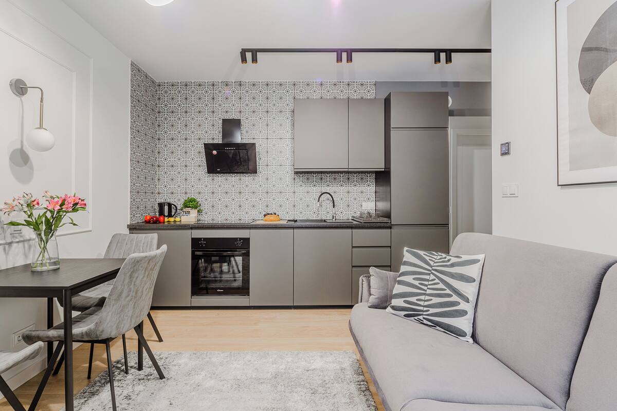 Eine kompakte Küche mit modernen Geräten, ein Essbereich mit Tisch und Stühlen sowie ein graues Sofa mit einem Zierkissen.