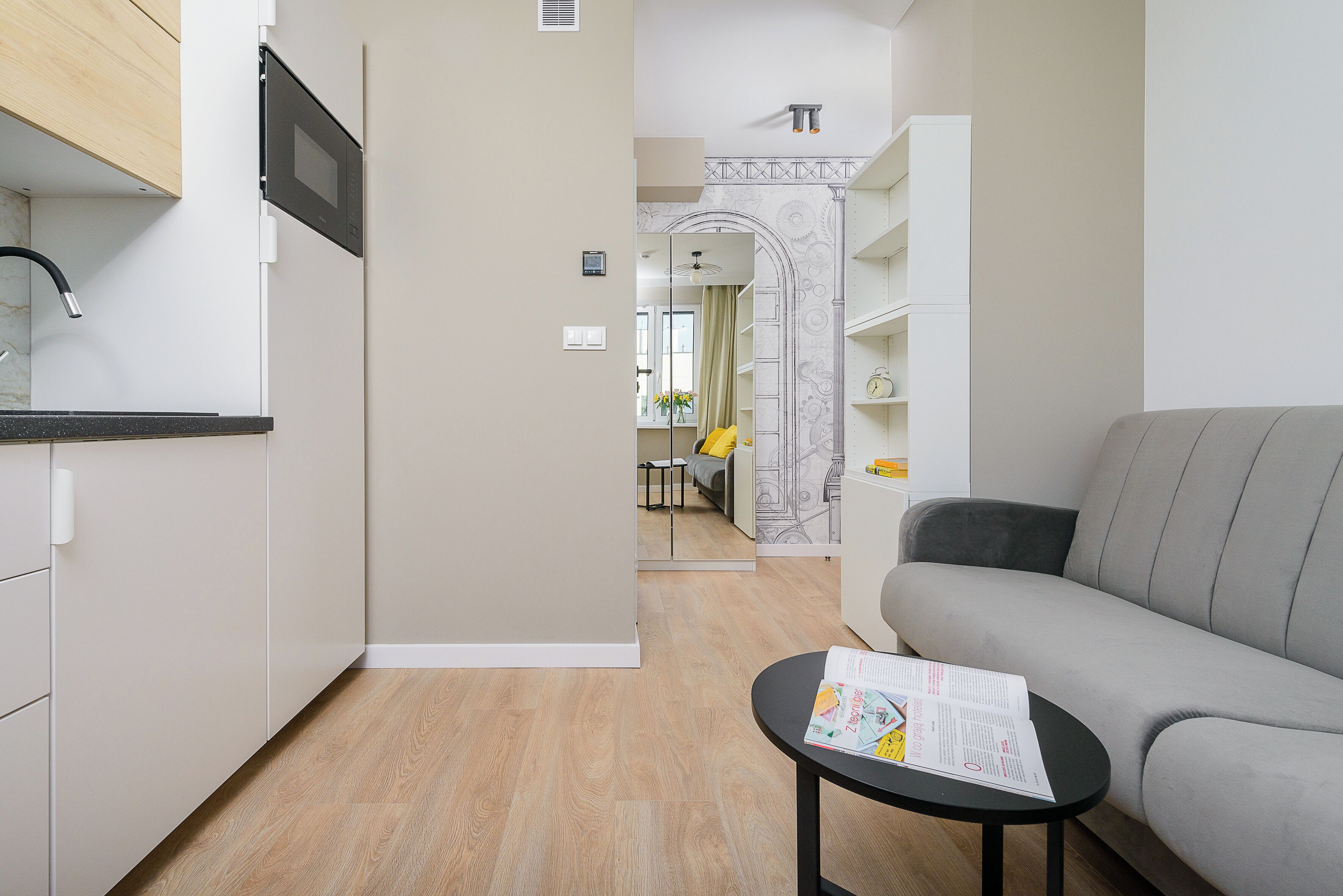 A modern living room with a grey sofa, a small black table with a magazine, and a kitchen area with a microwave and cabinets.
