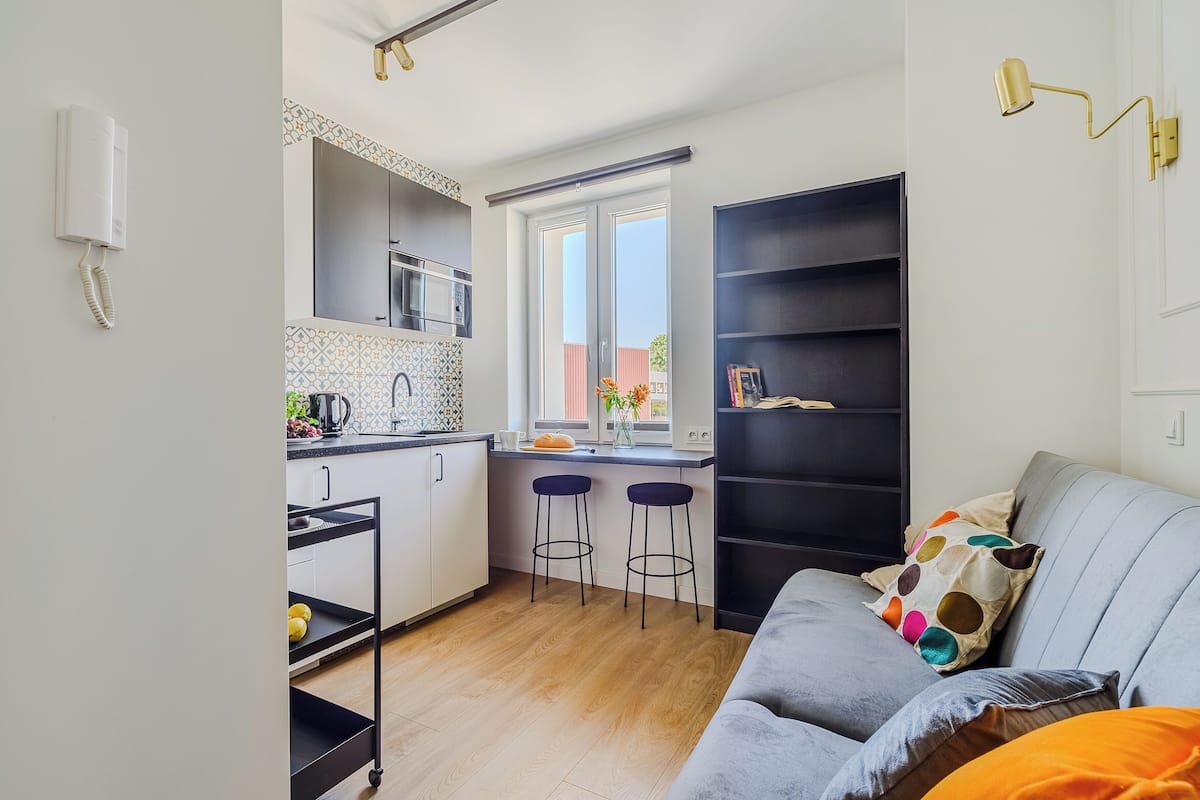 Ein kompakter Wohnraum mit Küchenzeile, einem grauen Sofa mit orangenen Kissen und einem schwarzen Bücherregal.