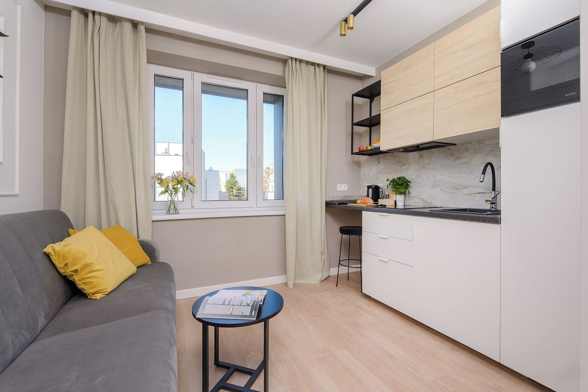 Eine moderne Wohnung mit einem grauen Sofa, einem kleinen runden Tisch, einer Küche mit weißen Schränken, einer schwarzen Arbeitsplatte und einem Fenster mit Vorhängen.