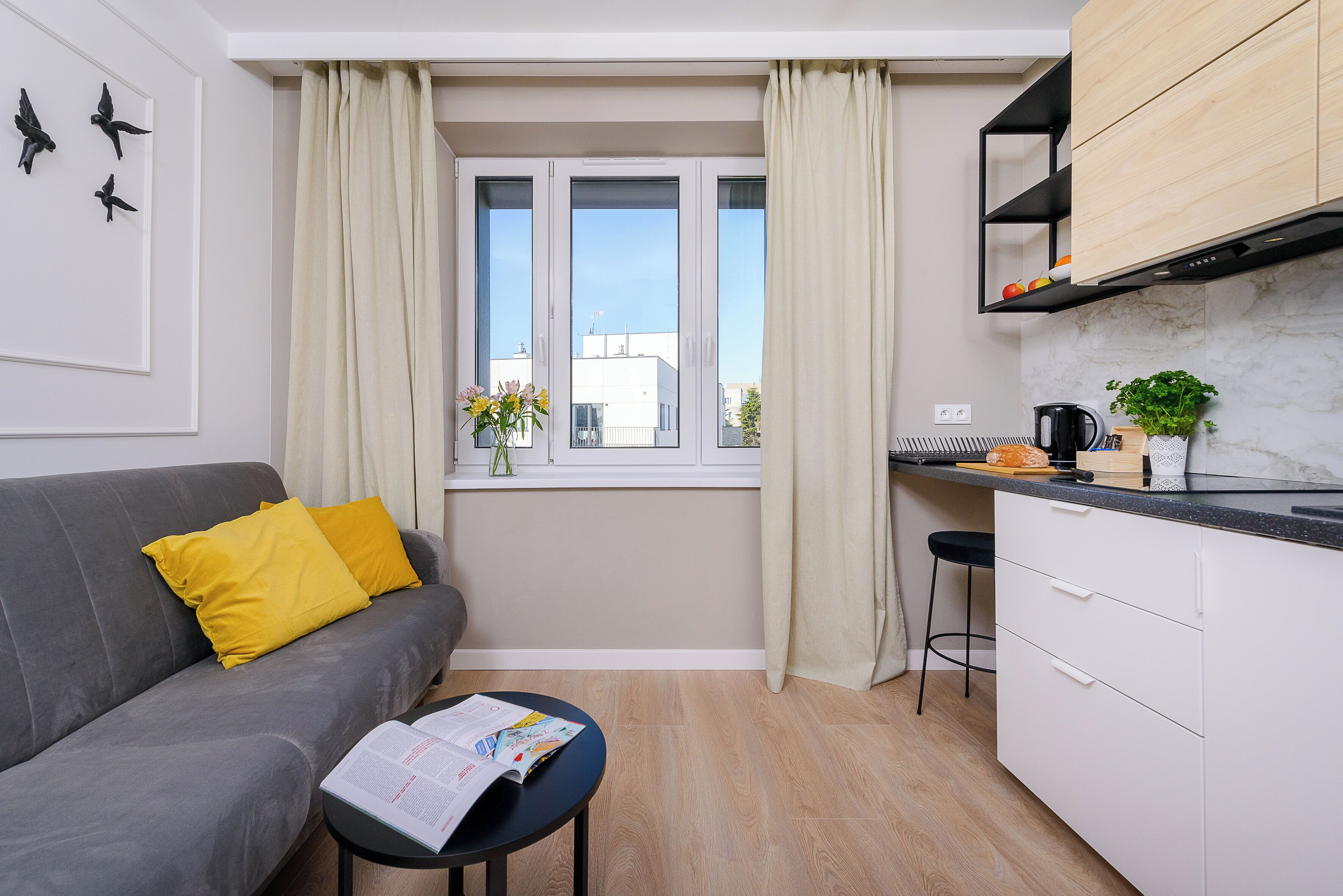Un espacio habitable pequeño con un sofá gris, almohadas amarillas, una mesita redonda con una revista, una ventana con cortinas, una zona de cocina con encimera negra y un estante de madera sobre el fregadero.
