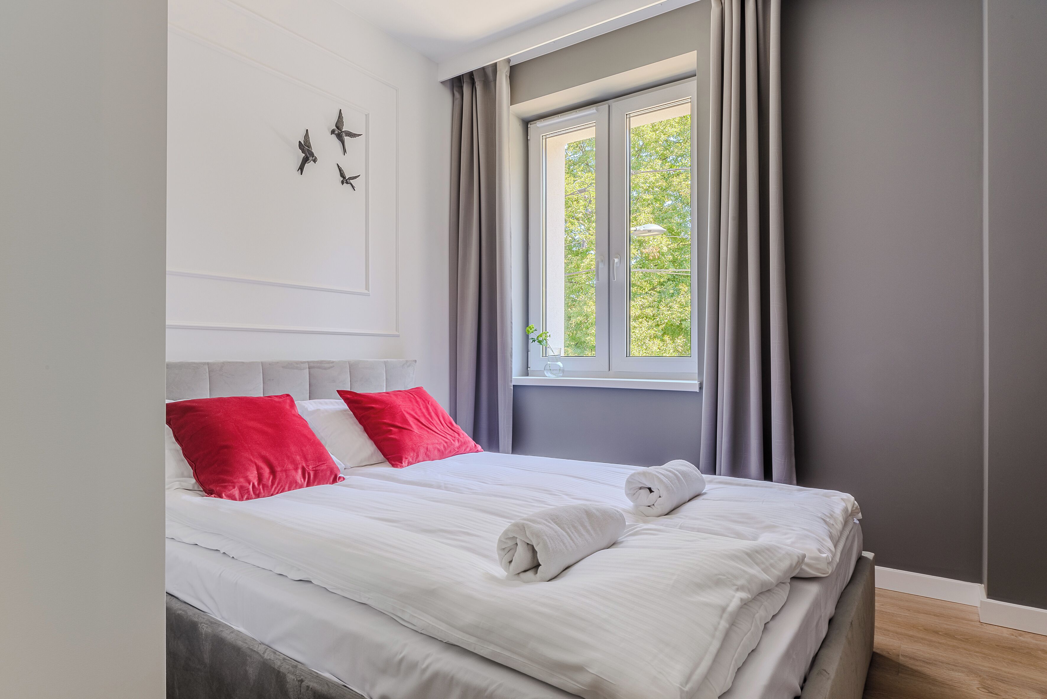 A neatly made bed with white and red pillows, a window with curtains, and a wall with a bird decoration.