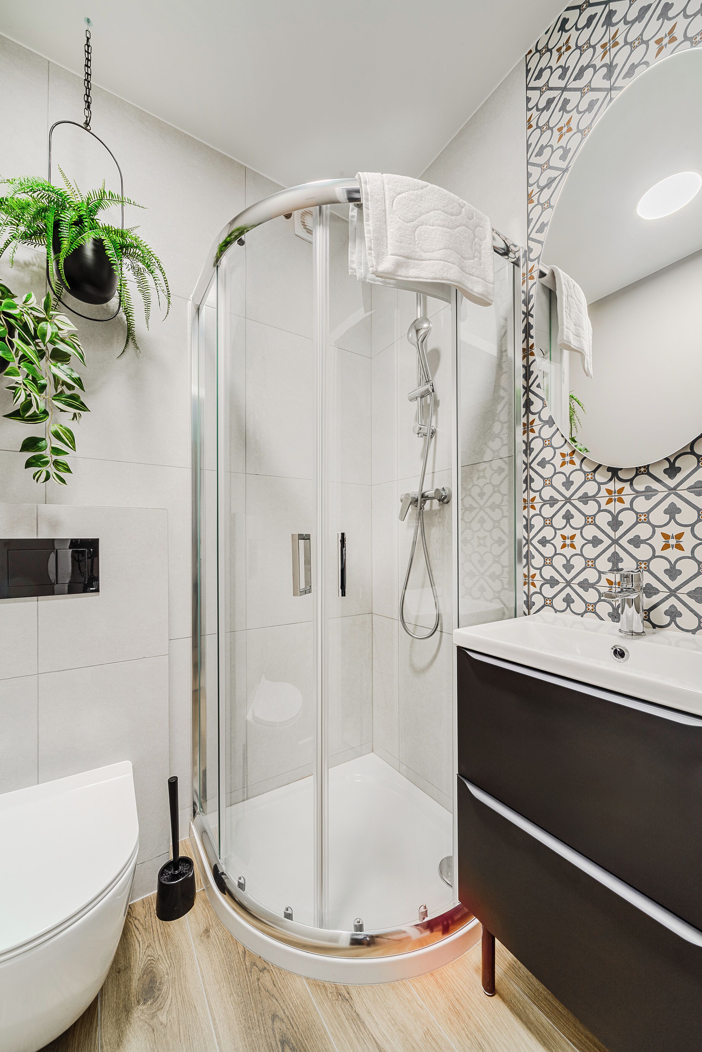 Une salle de bain moderne avec une douche à l’italienne en verre, des toilettes blanches, un meuble-lavabo noir et un mur recouvert de carreaux décoratifs.