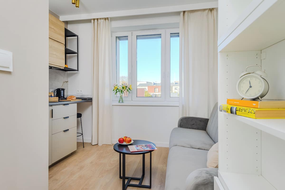 Ein kompakter Wohnraum mit Küchenzeile, einer Couch, einem kleinen Tisch mit einer Zeitschrift und einem Fenster mit Vorhängen.
