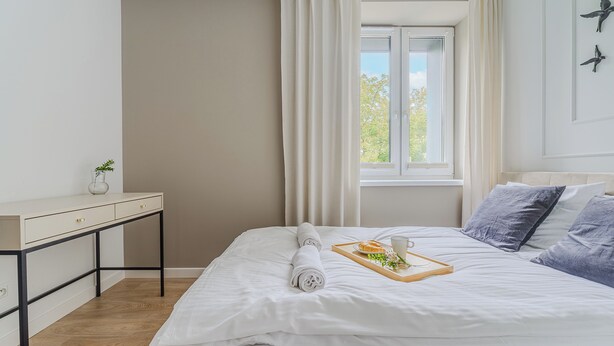 Una cama bien hecha con una toalla enrollada, una bandeja con comida y un vaso, y un escritorio con una planta.