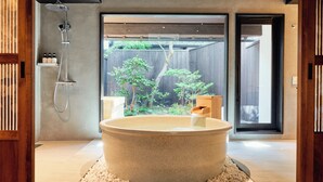 A modern bathroom with a large stone bathtub, a shower, and a view of a wooden deck and trees outside. - Kyoto Machiya Kamogawa Gardens (Kyoto)
