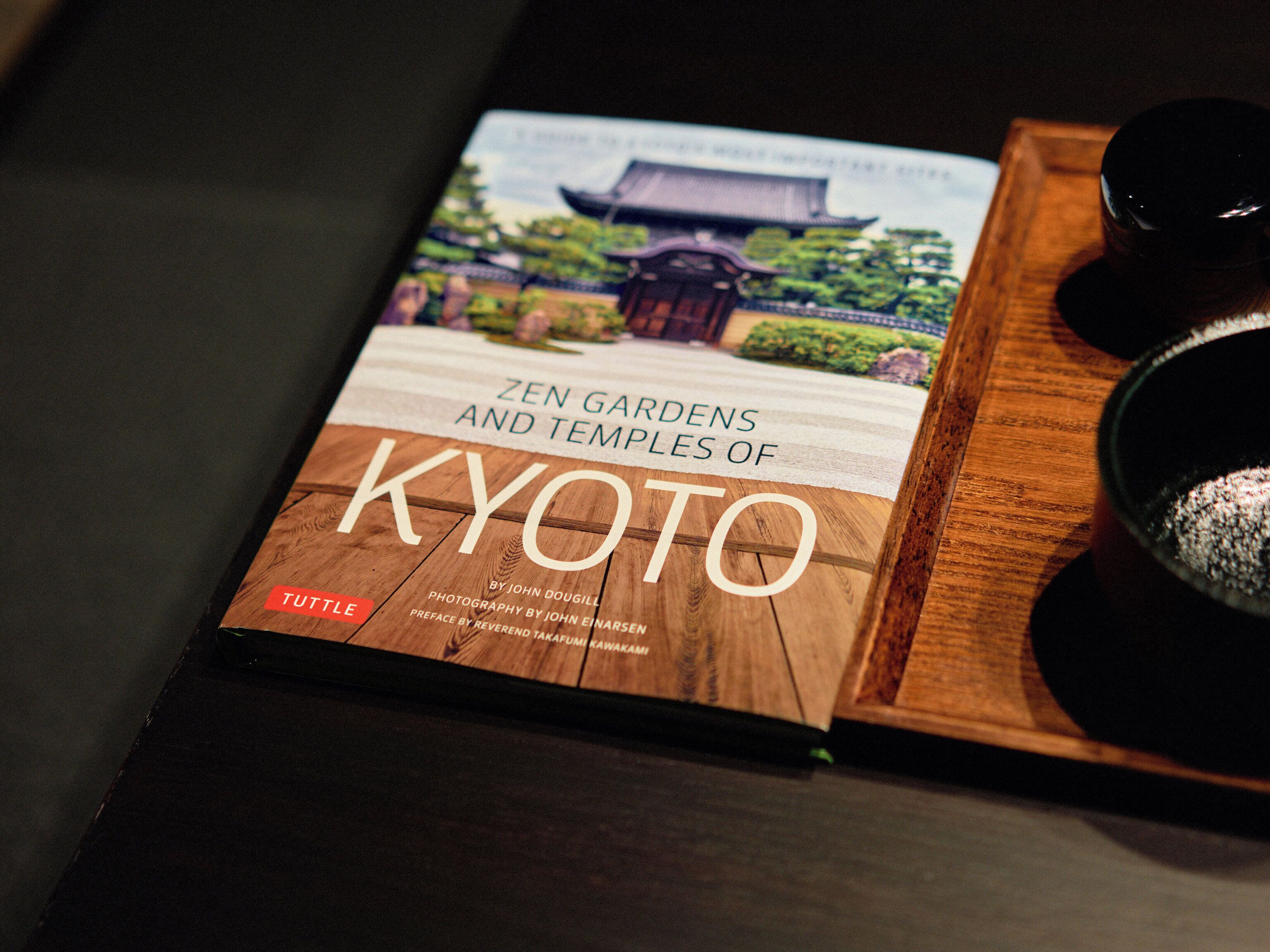 A book titled "Zen Gardens and Temples of Kyoto" by John Dougill, with a wooden tray and two black cups beside it.