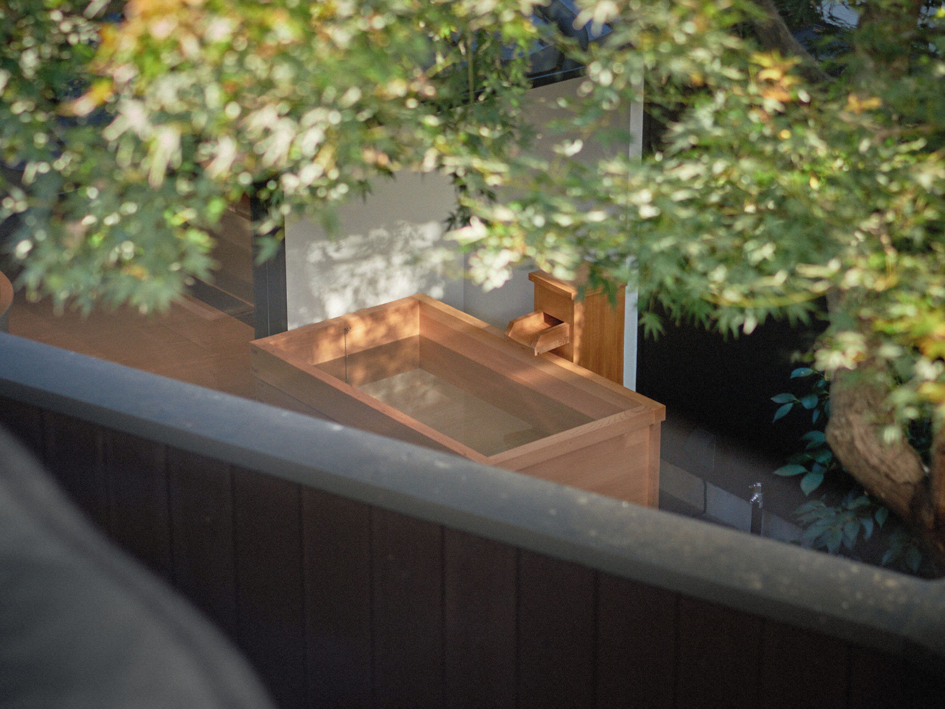 A wooden bathtub with a small wooden bench, surrounded by greenery and a white wall.
