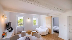 A modern living room with a wooden floor, white walls, and a white sofa. There are two windows providing natural light, a desk with a computer, and a dining table with chairs.