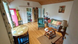 Een gezellige woonkamer met een houten vloer, een groene fauteuil, een bank met kleurrijke kussens, een salontafel met boeken en een afstandsbediening, en een eettafel met stoelen.
